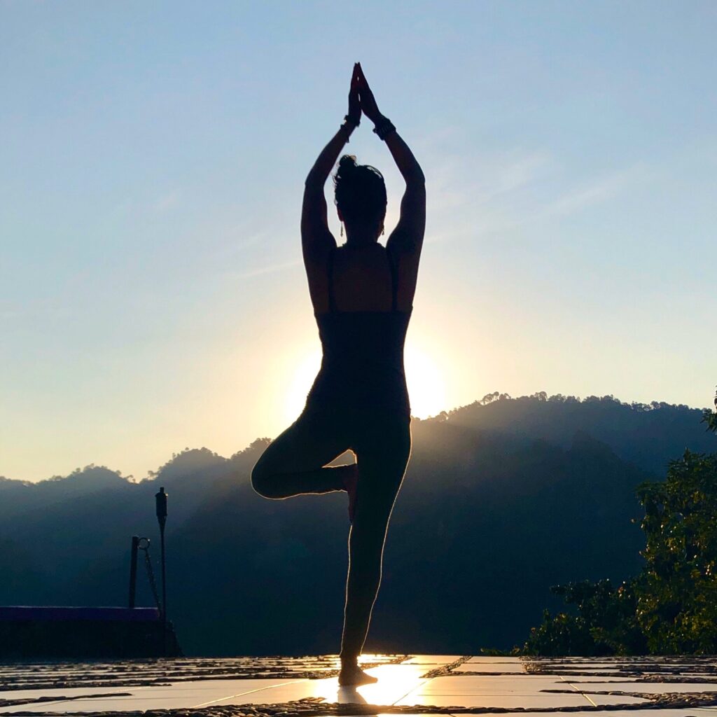 Frau vor der Sonne stehend bei der Ausführung der Yoga-Übung "Baum"